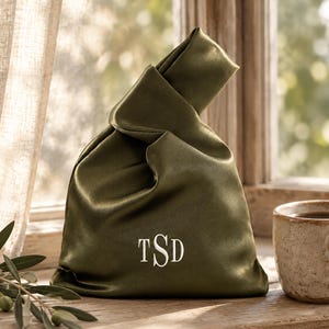 May include: A dark olive green satin knot bag featuring a smooth, lustrous finish and a single handle formed by the fabric. The front of the bag is embroidered with the white monogram letters TSD in a classic serif font, centered near the base.