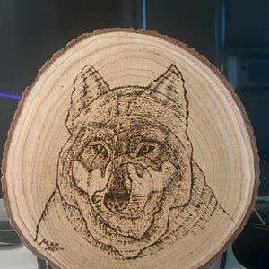 May include: A circular wood slice featuring a pyrography illustration of a wolf's face. The natural light-colored wood grain is visible, with a dark brown bark edge. The wolf is rendered in detailed black burn marks, with a signature in the corner.