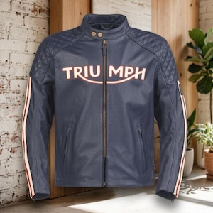May include: A navy blue perforated leather motorcycle jacket featuring the word TRIUMPH in white and red lettering across the chest. The jacket includes a mandarin collar with two snap buttons, quilted shoulder panels, and vertical side stripes.