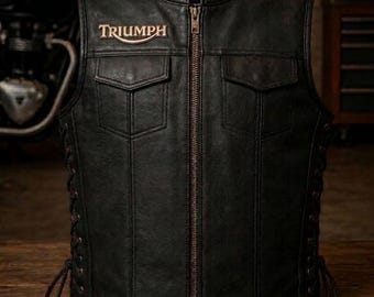 May include: A black leather vest featuring a central metal zipper, two chest pockets with flaps, and side lacing details. The word Triumph is embroidered in a light color on the upper right chest area of the garment, which has a round neckline.