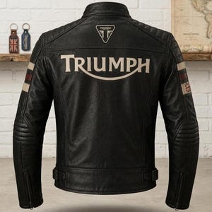 May include: A black leather motorcycle jacket featuring the word TRIUMPH in large beige lettering across the back. The jacket includes a small triangular logo at the neck, quilted shoulder panels, and sleeve stripes with a Union Jack flag detail.