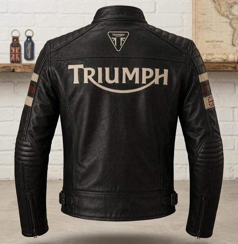 May include: A black leather motorcycle jacket featuring the word TRIUMPH in large beige lettering across the back. The jacket includes a small triangular logo at the neck, quilted shoulder panels, and sleeve stripes with a Union Jack flag detail.