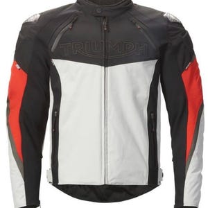 Può includere: Una giacca da moto Triumph nei colori nero, bianco e rosso. La giacca presenta un colletto alto, una cerniera frontale a tutta lunghezza e tasche con zip sul petto e sui lati. La parola TRIUMPH è impressa sul petto. Le maniche hanno inserti rossi.