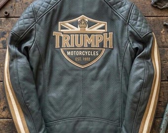 May include: A dark gray leather motorcycle jacket featuring a perforated texture, quilted shoulder panels, and light brown racing stripes on the sleeves. The back displays a large Triumph Motorcycles logo with a crown and flag graphic, EST. 1902.