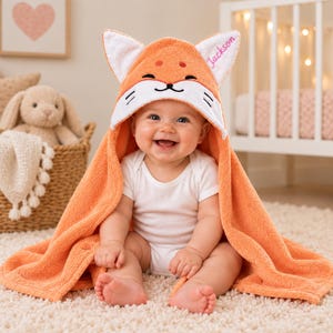 May include: An orange hooded towel featuring a fox face design with white accents and pointed ears. The name Jackson is embroidered in pink script on one ear. The towel is made of soft, textured fabric and is draped over a smiling infant.