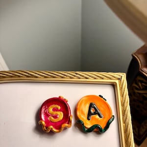May include: Two small, circular, handmade clay trinket dishes. The left dish is magenta with a gold letter S and gold trim. The right dish is orange with a black letter A and dark green trim. Both have a glossy, uneven, organic finish.