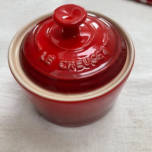 May include: A round, cherry red stoneware ramekin with a matching lid. The lid features a central knob handle and the embossed text LE CREUSET. The interior rim and the underside of the lid are finished in a contrasting light beige glaze.