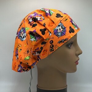 May include: An orange Halloween-themed scrub cap featuring a repeating pattern of Mickey Mouse, Minnie Mouse, spiderwebs, and jack-o'-lanterns. The cap has a drawstring and is displayed on a mannequin head.