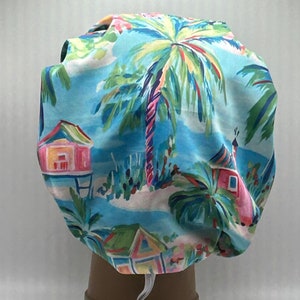 May include: A blue and white fabric shower cap with a tropical print featuring palm trees, pink and green houses, and white sand. The shower cap has a white drawstring.