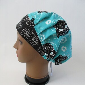 May include: A turquoise and black scrub hat with a white Star Wars logo and a black Darth Vader skull design. The hat has a white drawstring.
