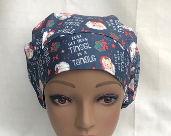 May include: A dark blue scrub cap with a repeating pattern of Santa Claus faces, wrapped gifts, and stars. The text "Don't Get Your Tinsel in a Tangle" is printed on the cap. The cap is displayed on a mannequin head.