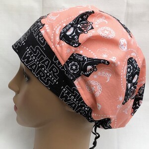 May include: A pink and black scrub hat with a Star Wars theme. The hat features a repeating pattern of Darth Vader's helmet with a sugar skull design. The text "Star Wars The Force Awakens" is printed on the black band around the hat.