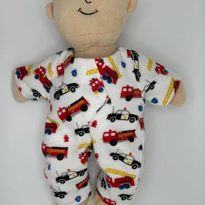 K&ouml;nnte beinhalten: Eine weiche, beige Puppe, die einen wei&szlig;en Strampler mit roten Feuerwehrautos, schwarzen Polizeiautos und gelben Hydranten tr&auml;gt. Die Puppe hat ein l&auml;chelndes Gesicht und ein kleines gelbes Haaraccessoire.