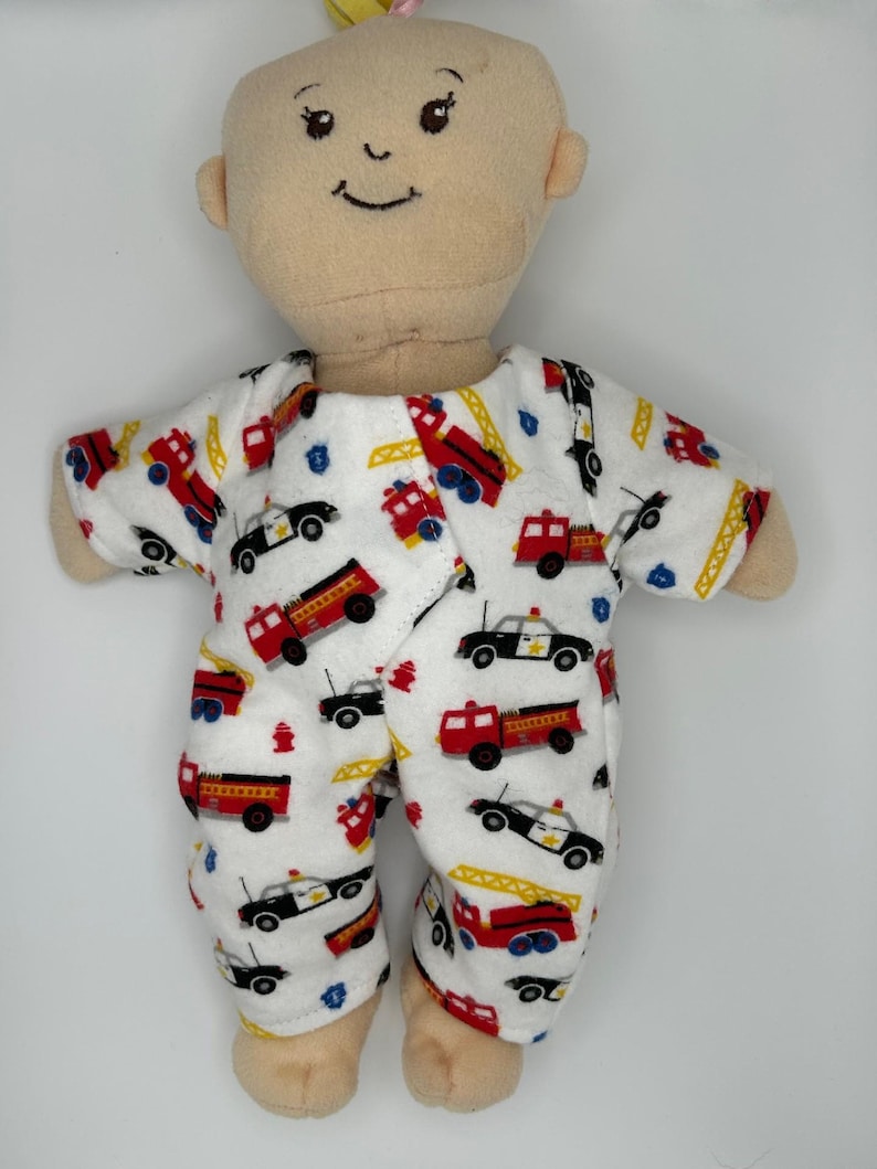 K&ouml;nnte beinhalten: Eine weiche, beige Puppe, die einen wei&szlig;en Strampler mit roten Feuerwehrautos, schwarzen Polizeiautos und gelben Hydranten tr&auml;gt. Die Puppe hat ein l&auml;chelndes Gesicht und ein kleines gelbes Haaraccessoire.