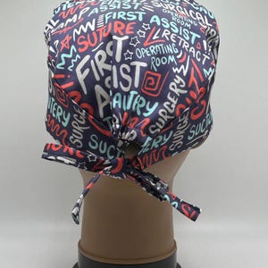 May include: A navy blue surgical scrub cap with a colorful graffiti-style design. The cap features words like "First", "Assist", "Surgery", and "Operating Room" in white, red, and teal. It is tied at the back with a matching bow.