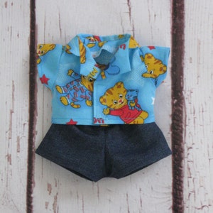 May include: A blue and white patterned shirt with a red collar and red stars. The shirt features a cartoon tiger wearing a red shirt and holding a small tiger. The shirt is paired with blue denim shorts.