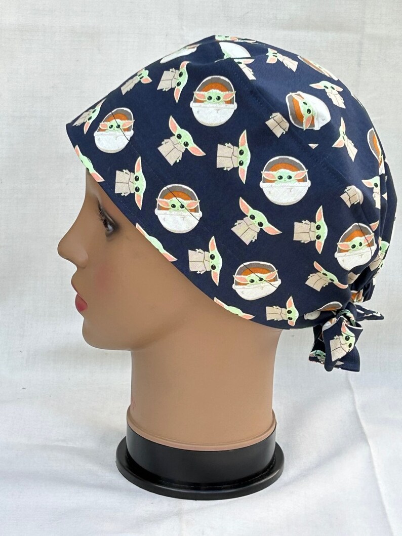 May include: A blue surgical scrub hat with a repeating pattern of a green alien character in a pod. The hat has a tie at the back.