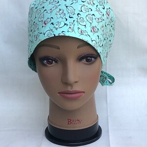 May include: A turquoise blue surgical scrub hat with a repeating pattern of cartoon genie faces. The hat has a tie on the back.