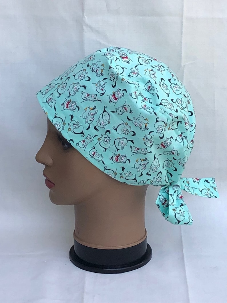 May include: A turquoise blue scrub hat with a repeating pattern of cartoon genie characters. The hat has a tie at the back.