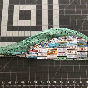May include: A green scrub hat with a pattern of license plates from different states. The license plates have various slogans and sayings, such as "I Luv U", "2 Cool 4U", and "URA QT 2".