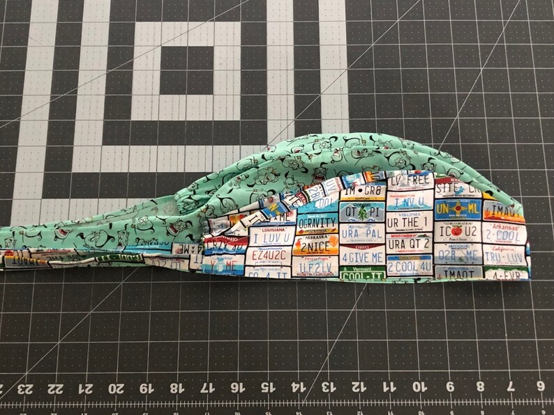 May include: A green scrub hat with a pattern of license plates from different states. The license plates have various slogans and sayings, such as "I Luv U", "2 Cool 4U", and "URA QT 2".