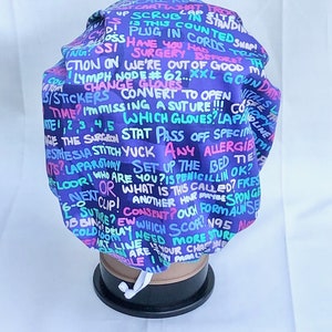 Peut inclure: Un bonnet de scrub chirurgical violet avec un imprimé coloré de termes et de phrases médicaux. Le bonnet a un cordon de serrage blanc.