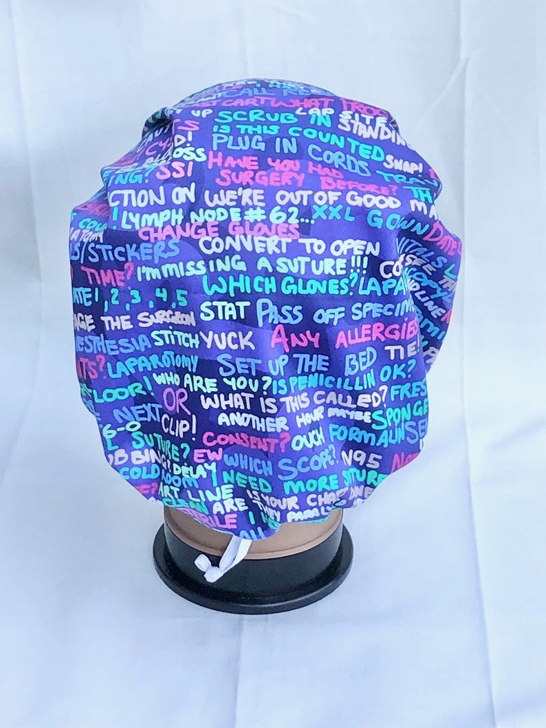 May include: A purple surgical scrub hat with a colourful print of medical terms and phrases. The hat has a white drawstring.