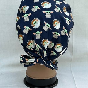 May include: A dark blue scrub hat with a repeating pattern of a green alien character in a pod. The hat has a tie at the back.