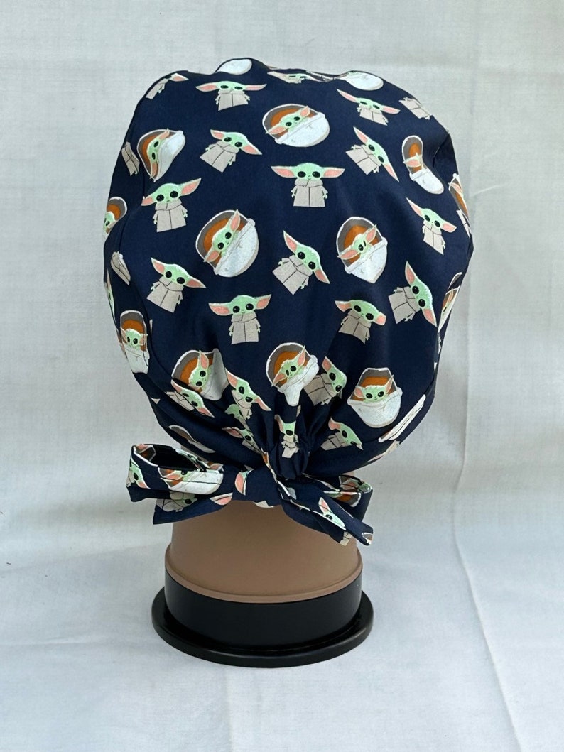 May include: A dark blue scrub hat with a repeating pattern of a green alien character in a pod. The hat has a tie at the back.