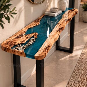 Op de afbeelding: Een sidetable met een natuurlijke houten rand en een centrale rivier van doorschijnende blauwe epoxyhars. Het harsgedeelte bevat ingebedde gladde grijze rivierstenen. De tafel wordt ondersteund door twee rechthoekige zwarte metalen poten.