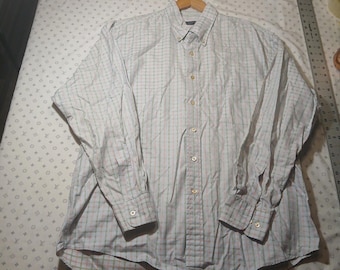 May include: A long-sleeve button-down shirt featuring a light-colored fabric with a subtle grid pattern in soft green and pink tones. The shirt includes a classic collar, a front button placket, and buttoned cuffs, displayed next to a metal ruler.