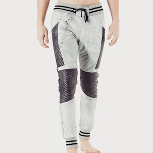 May include: Light gray joggers featuring a distressed, mottled texture with black ribbed panels on the thighs and knees. These pants include a black drawstring waistband, black striped cuffs at the ankles, and a slim-fit design throughout.