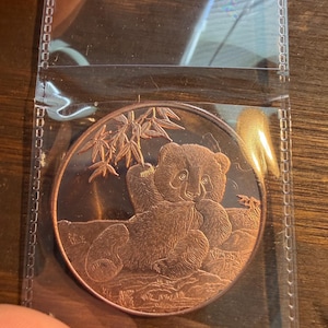 May include: A round copper-colored coin featuring a detailed relief of a panda sitting and eating bamboo stalks. The panda is positioned in the center with bamboo leaves visible above its head. The coin is encased in a clear plastic sleeve.