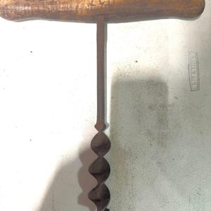 May include: A vintage T-handle hand auger drill with a weathered wooden crossbar featuring the carved letters AB. The tool has a long, straight metal shaft connected to a dark, rusted spiral drill bit at the bottom end for boring holes.