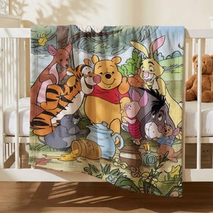 Op de afbeelding: Een zachte babydeken met een kleurrijke illustratie van Winnie de Poeh, Teigetje, Knorretje, Iejoor, Konijn, Kanga, Roe en Gopher in het Honderd Bunderbos. De afbeelding bevat een honingpot met het label HUNN en een blauwe theepot.
