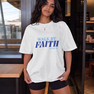May include: A white short-sleeved t-shirt featuring the blue text WALK BY FAITH and the smaller blue text 2 COR 5:7 printed on the chest. The shirt has a crew neckline and a relaxed fit, paired with black shorts.