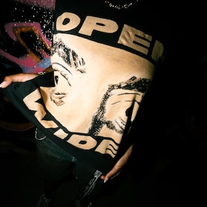 May include: A black short-sleeved t-shirt featuring a large tan graphic print of two faces with eyes looking in different directions. The text "OPEN" is printed in bold black letters at the top, and "WIDE" is printed in black letters at the bottom.