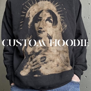 Puede incluir: Una sudadera con capucha negra que presenta un gran estampado gráfico beige desgastado de una figura con halo en la parte delantera. El texto CUSTOM HOODIE está impreso en blanco en el centro del pecho. La prenda tiene mangas largas y un bolsillo.