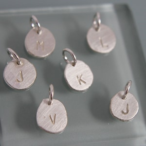 May include: Six silver charms with different letters on them. The letters are J, M, K, V, and two Js.