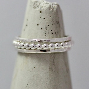 May include: A close-up of three silver rings. The top and bottom rings have a hammered texture, while the middle ring is adorned with small, evenly spaced silver beads. The rings are displayed on a gray, cone-shaped object.