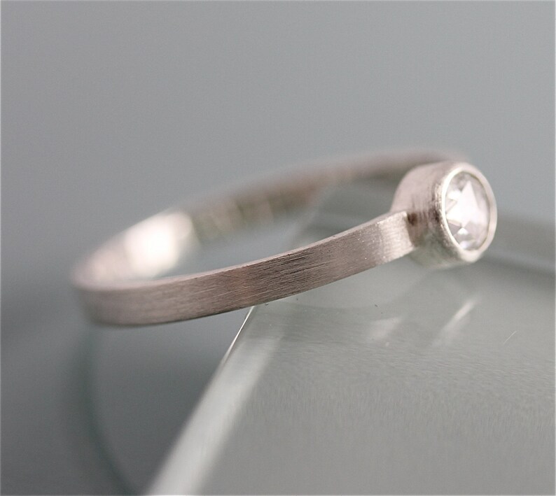 May include: A simple silver band ring with a single, small, round, clear gemstone set in a bezel setting.