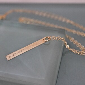 May include: A gold necklace with a thin chain and a long, rectangular pendant with an inscription. The pendant is made of brushed metal and has a slightly textured surface.