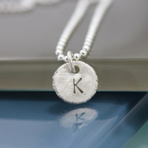 May include: A silver necklace with a round pendant featuring the letter K. The pendant has a textured surface and a simple design.
