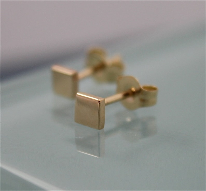May include: A pair of gold square stud earrings. The earrings are small and simple, with a polished finish.