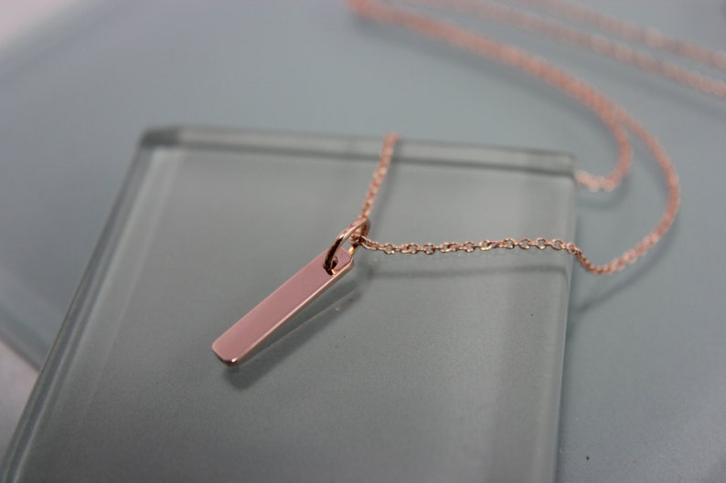 Puede incluir: Un collar de oro rosa con una cadena fina y un colgante rectangular largo.