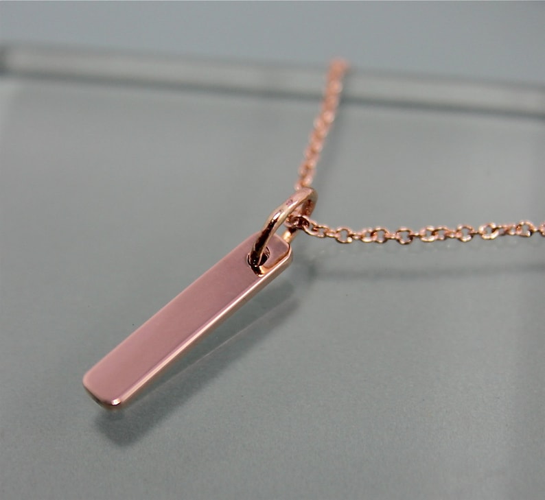 Puede incluir: Un collar de oro rosa con un colgante rectangular simple. El colgante es liso y pulido, y la cadena es delicada y fina.
