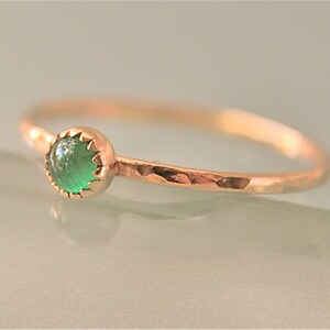 May include: A delicate gold ring featuring a round, vibrant green gemstone. The ring has a hammered texture on the band and a scalloped bezel setting. The gemstone is the focal point, set against the warm gold.