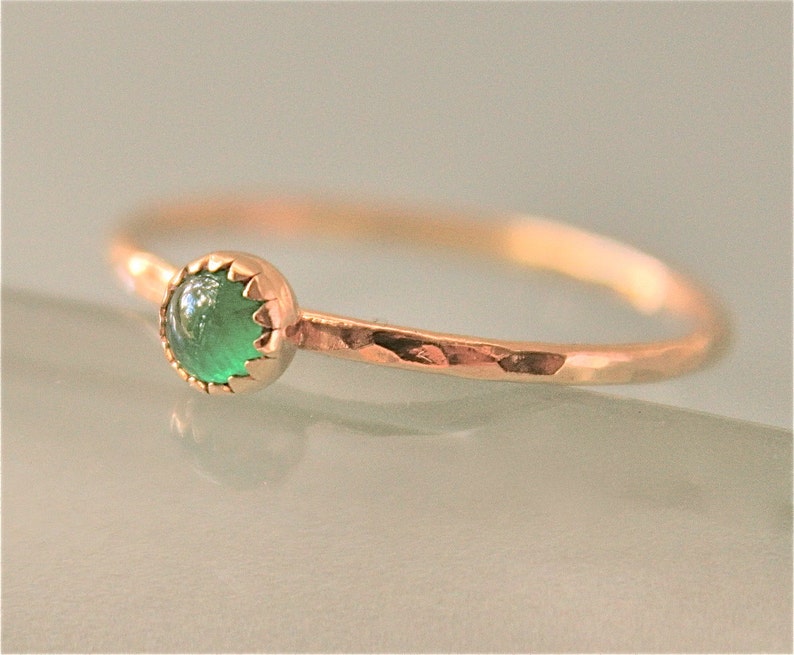May include: A delicate gold ring featuring a round, vibrant green gemstone. The ring has a hammered texture on the band and a scalloped bezel setting. The gemstone is the focal point, set against the warm gold.