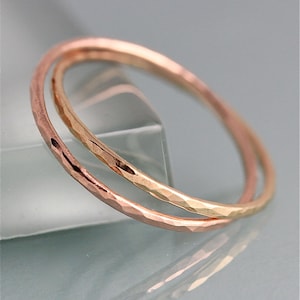 May include: Two hammered metal bangles, one rose gold and one yellow gold, are intertwined. The bangles have a textured surface and a simple, elegant design. The jewelry is displayed on a reflective surface.