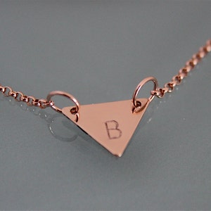 May include: A rose gold necklace with a triangle pendant featuring the letter "B" engraved on it.